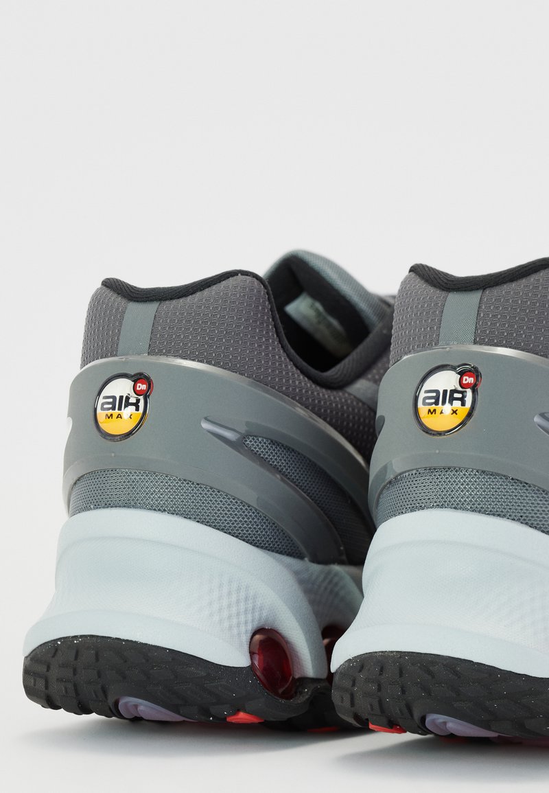 Zapatilla gris con materiales de malla y goma, con un talón redondeado, amortiguación de aire visible y acentos de logotipo en amarillo y negro.