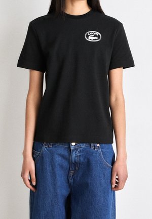 Kvinde iført en ensfarvet sort Lacoste t-shirt med hvidt logo på brystet og løse blå jeans mod en ensfarvet baggrund.
