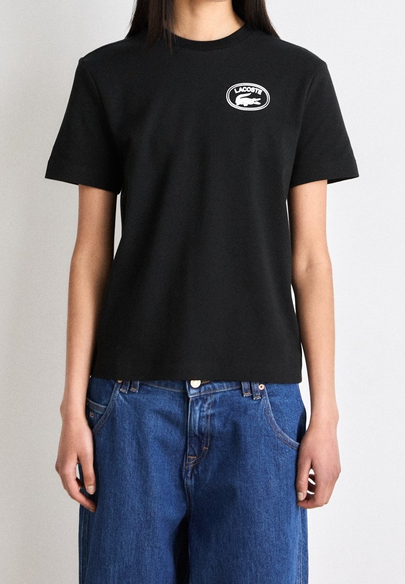 Femme portant un t-shirt Lacoste noir uni avec un logo blanc sur la poitrine et un jean bleu large sur un fond uni.