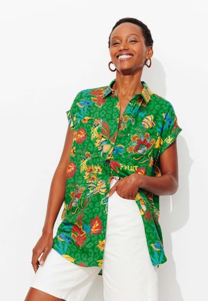 Femme souriante aux cheveux courts portant des boucles d'oreilles créoles en or, une chemise à fleurs verte éclatante boutonnée et un short blanc, main dans la poche.