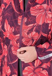 Ruka zapíná fialovou bundu s velkými růžovými a červenými květinovými vzory a reflexní nálepkou "HYRAR" na rukávu.