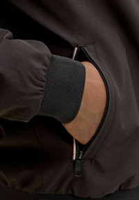 Χέρι εν μέρει μέσα στην τσέπη με φερμουάρ ενός μαύρου μπουφάν με ριμπ μανσέτα και λεπτομέρεια ριγέ επένδυσης στο φερμουάρ.