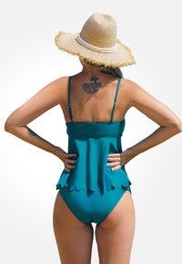 Costume da bagno tankini turchese con orlo scallop, spalline sottili e un tatuaggio floreale visibile sulla schiena. Indossato con un cappello di paglia a tesa larga.