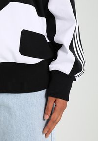 Schwarz-weißes Sweatshirt mit markantem Grafikdesign, gerippten Bündchen und drei weißen Streifen an den Ärmeln. Darunter sind leichte Jeans aus Denim sichtbar.