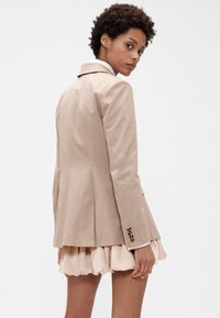 Bež sakoi s uklopljenim krojem, reverom s urezima, dugim rukavima i pet dugmadi na manžetama, upareni s laganom suknjom s naborima.