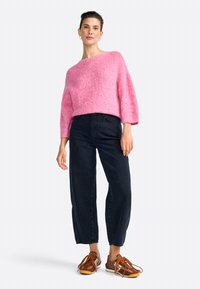 Maglione rosa in maglia morbida con spalle scese, abbinato a jeans in denim neri a vita alta e sneakers marroni con lacci arancioni.