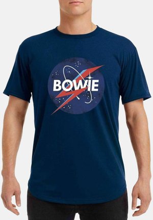 Uomo che indossa una t-shirt blu scuro con un disegno circolare a tema spaziale, un fulmine rosso e la parola "BOWIE" in lettere bianche.