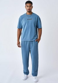 Camiseta de algodón azul claro con mangas cortas y un logo negro en el pecho, combinada con pantalones de chándal azules claros a juego y zapatillas blancas.