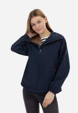 Jeune femme aux longs cheveux portant une veste zippée bleu marine sur une chemise à rayures, debout avec une main derrière la tête.