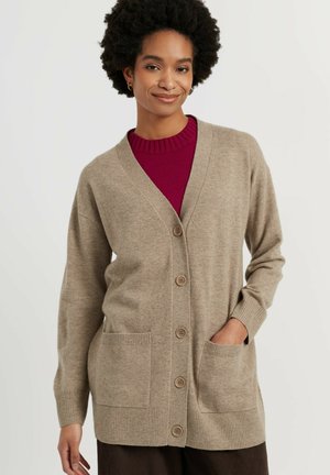 Vrouw draagt een beige vest met knopen over een rode trui, glimlachend met één hand in de zak van het vest tegen een effen achtergrond.