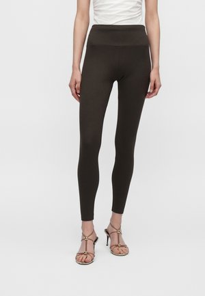 Vrouw met donkerbruine legging met hoge taille en open sandalen met hoge hakken tegen een effen lichte achtergrond.