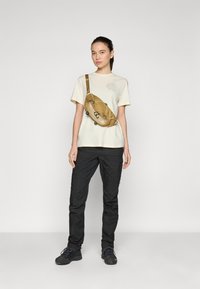 Svijetlo bež majica s kružnim logotipom, crne cargo hlače i bež fanny pack s patentnim zatvaračima i detaljima od užeta, nošena preko ramena.