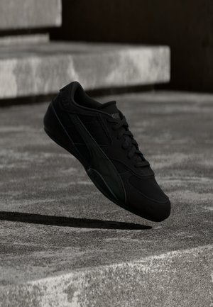Chaussure de sport noire à lacets, flottant au-dessus de marches en béton dans une lumière naturelle douce avec des ombres marquées.