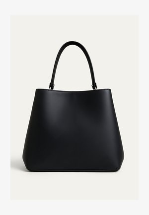 Femme aux longs cheveux noirs portant un costume vert sauge, tenant un grand sac à main noir, avec un collier et des boucles d'oreilles argentés volumineux, posant devant un mur texturé.