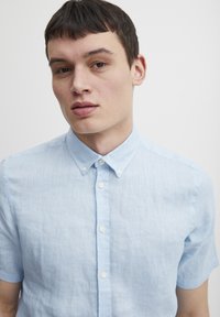 Camisa de lino azul claro de manga corta con cuello abotonado, que presenta botones blancos y un sutil bordado en el pecho.
