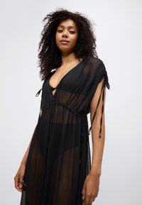 Μαύρο διαφανές φόρεμα με βαθύ V-λαιμόκοψη, ζώνη με κορδόνι στη μέση και κορδόνια στα μανίκια. Ελαφρύ ύφασμα με ρέουσα σχεδίαση.