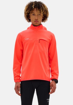Giovane con capelli ricci che indossa una maglia sportiva corallo brillante con zip e maniche lunghe e leggings neri, con un orologio fitness al polso sinistro.