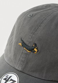Casquette de baseball grise en coton avec une texture côtelée. Présente un design brodé de teckel en noir et jaune ainsi qu'une étiquette ronde avec logo.