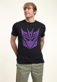 Zwart T-shirt met korte mouwen met een groot paarse grafisch ontwerp van een gestileerd embleem aan de voorkant. Gladde stofstructuur.