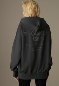 Yliotettu musta huppari, jossa on teksturoitu printti, jossa lukee "BROOKLYN 1995" ja "NEW YORK CITY" metallisilla yksityiskohdilla takana.