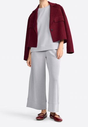 Femme portant un pantalon ample gris clair et un haut avec une veste courte rouge foncé drapée sur les épaules et des chaussures plates rouges assorties.