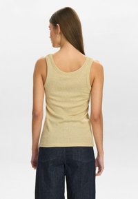 Fitted gold tank top with a scooped back, textured fabric, and wide shoulder straps. Paired with dark denim trousers, showing a casual style.