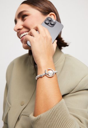 Pulsera de plata con cuentas esféricas y un acento de perla, llevada en una muñeca mientras sostiene un teléfono inteligente. Sudadera con una manga texturizada visible.