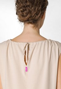 Beige bezrukávový top s širokým výstřihem, uzlovým detailem na zadní straně a malou růžovou visačkou. Hladká tkanina s jemnou texturou.