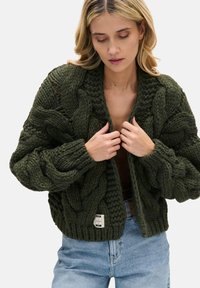 Gruby, oliwkowozielony kardigan dziergany z wzorem warkocza, z otwartym przodem i ściągaczem u dołu. Posiada opuszczane ramiona oraz teksturowane detale na rękawach.