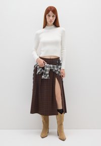 Witte geribde lange mouwen crop top gecombineerd met een bruine geblokte maxi rok met een voorkleur en gespsluiting, gedragen met tan enkellaarsjes.
