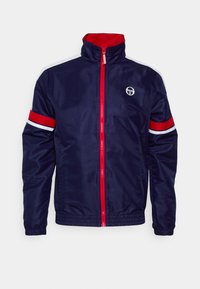 Marine vindjakke med høy krage, rød glidelås og kontrasterende rød og hvit stripete detaljer på ermene. Har logo på brystet.