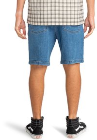 Achteraanzicht van een persoon in blauwe denim shorts, een beige geruite blouse en zwarte hoge sneakers met witte zolen op een witte achtergrond.