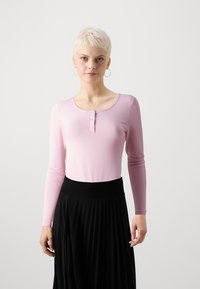Langærmet pink bomuldsbluse med en rund udskæring og tre knaplukninger, parret med en sort plisseret nederdel med en strækbare talje.