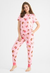 Ung kvinna som ler, står barfota i en rosa pyjamas med mönster av igelkottar och hjärtan mot en vit bakgrund.