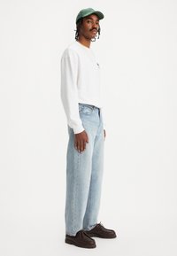 Witte longsleeve shirt, lichtblauwe rechte jeans, donkerbruine schoenen en een groene pet. Eenvoudig ontwerp met minimale accenten.