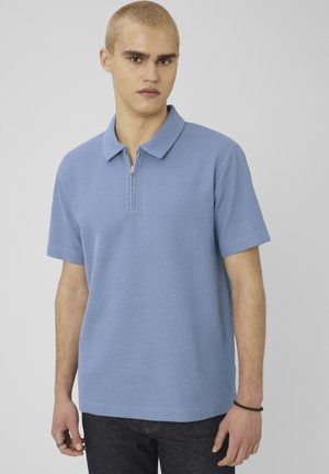 Jeune homme aux cheveux blonds courts portant un polo bleu clair à manches courtes avec une fermeture éclair à l'avant, debout devant un fond gris uni.