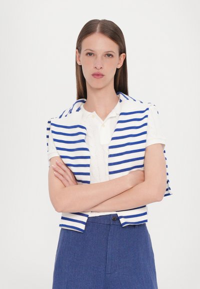 Un gilet court rayé bleu et blanc posé sur un polo blanc, associé à un pantalon bleu. Lignes épurées et tricot texturé dans un design décontracté.