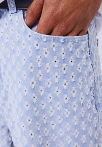 Pantalones cortos de mezclilla azul claro con un patrón texturizado de puntos, bordados blancos y detalles deshilachados cerca de los bolsillos.