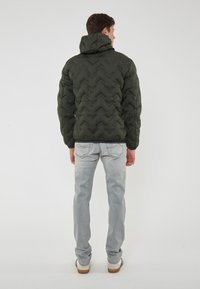 Giacca verde imbottita con cappuccio, caratterizzata da un motivo a zigzag e polsini a coste, abbinata a jeans azzurri chiari e sneaker bianche.