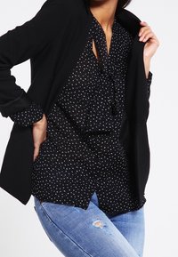 Schwarze Bluse mit weißen Punkten, mit einem Krawatten-Front und Rüschenausschnitt, kombiniert mit einem schwarzen Blazer und hellblauen, ausgewaschenen Jeans.