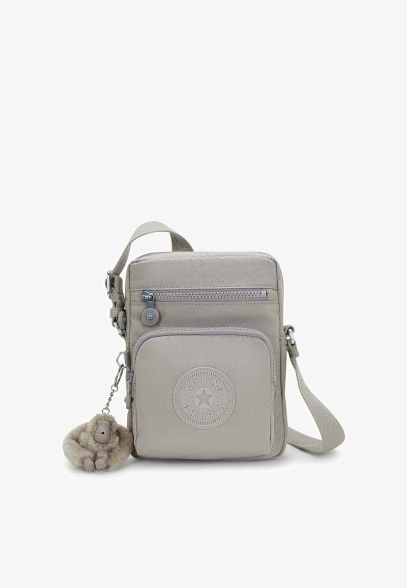 Beige crossbodytas gemaakt van lichtgewicht stof, met een ritssluiting, een rond logo en een pluchen apensleutelhanger eraan bevestigd.