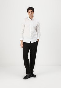 Weißes Hemd mit langen Ärmeln und schwarze Hose. Schwarze Lederschuhe vervollständigen das Outfit, das einen schmalen Schnitt und eine glatte Textur aufweist.