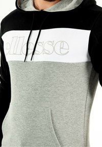 Sudadera con capucha en negro, blanco y gris. Presenta un gran logo frontal, cordones ajustables y una textura suave con un bolsillo tipo canguro.