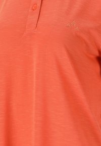 Korallfärgad polo med texturerad tyg, med en kort krage och en knapp. Litet logotypdetalj i guld på övre vänstra sidan.