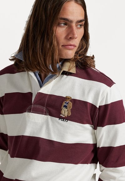 Un tânăr cu păr brunet lung până la umeri, îmbrăcat cu un tricou de rugby burgundy cu dungi albe, pe care se află brodat un ursuleț de pluș și textul "POLO".