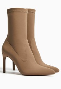 Vaaleanruskeat kangasnahkan nilkkurit tikku-koroilla, terävillä kärjillä ja saumattomalla muotoilulla. Sileä pinta, jossa yläosassa näkyvät tikkausyksityiskohdat.