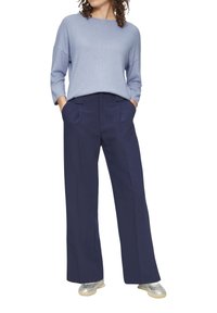Vrouw in een lichtblauwe langemouwen top, marineblauwe wijde broek met handen in de zakken en zilveren sneakers, staand tegen een witte achtergrond.
