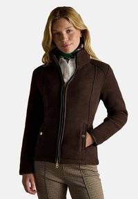 Kobieta w ciemnobrązowej kurtce na zamek, w kwiecistej szalu, białej koszuli i spódnicach w kratę houndstooth, stoi z jedną ręką w kieszeni.