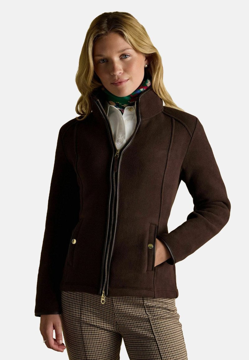 Kobieta w ciemnobrązowej kurtce na zamek, w kwiecistej szalu, białej koszuli i spódnicach w kratę houndstooth, stoi z jedną ręką w kieszeni.