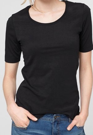 Kvinde iført en simpel sort T-shirt med korte ærmer og rund hals, stoppet ned i blå jeans, med hænderne i jeanslommerne.